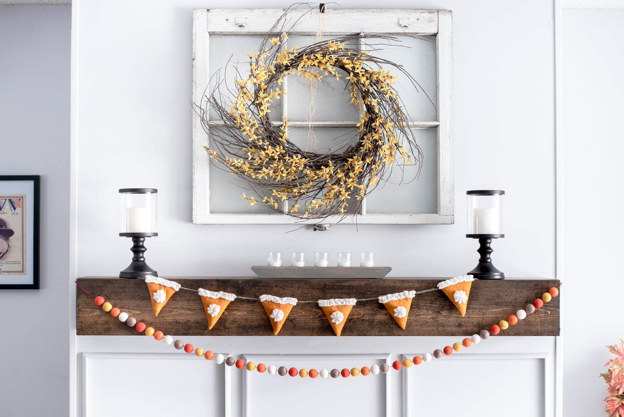 A mantel decorated with candy corn garland, a rustic wreath with yellow flowers on an old window frame, and two black candleholders with white candles.