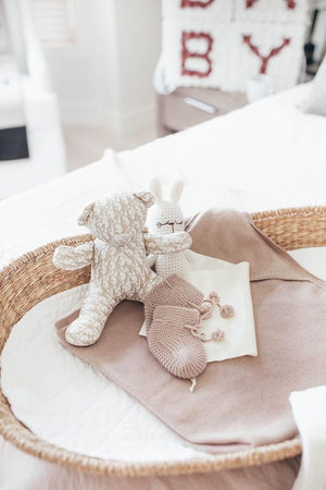 A woven basket holds a plush teddy bear, a knitted bunny, baby booties, and a folded cloth, set on a bed with a patchwork blanket.