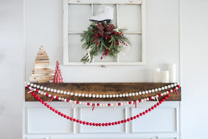 A holiday mantel decorated with red and white garlands, small Christmas trees, candles, and a wreath hanging on a window frame above.