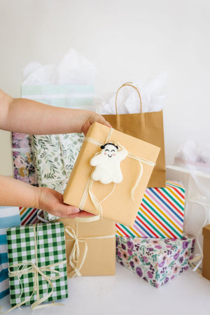 A person holds a brown paper-wrapped gift with a smiling ghost ornament, in front of a group of variously wrapped presents and gift bags.