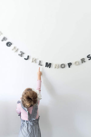 A child wearing a pink shirt and gray dress eagerly points at the Nivas Alphabet Garland (Gray) made of wool felt letters on a white wall.