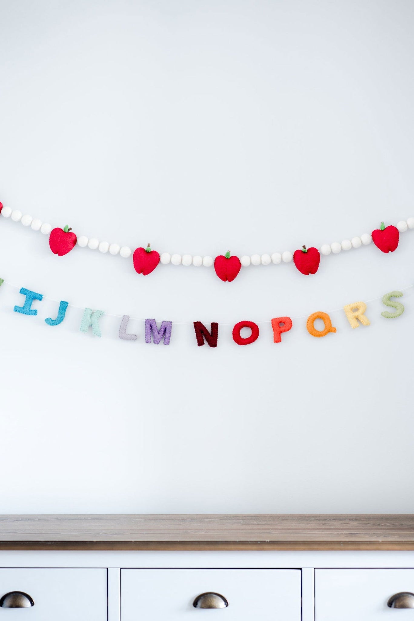 Nivass Apple Garland, featuring vibrant red apples and white wool beads, hangs alongside colorful wool felt alphabet letters, capturing the essence of the back-to-school season.