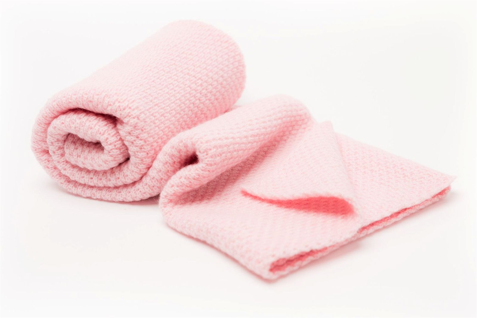 A rolled-up and partially unrolled pink baby blanket, the Nivas Cashmere Blend, crafted with soft wool, rests gracefully on a pristine white background.