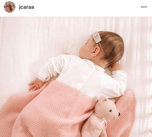 A baby sleeps on a bed covered with a soft pink Cashmere Blend Baby Blanket by Nivas, next to a pink stuffed teddy bear, wearing a white outfit with a small hair clip.