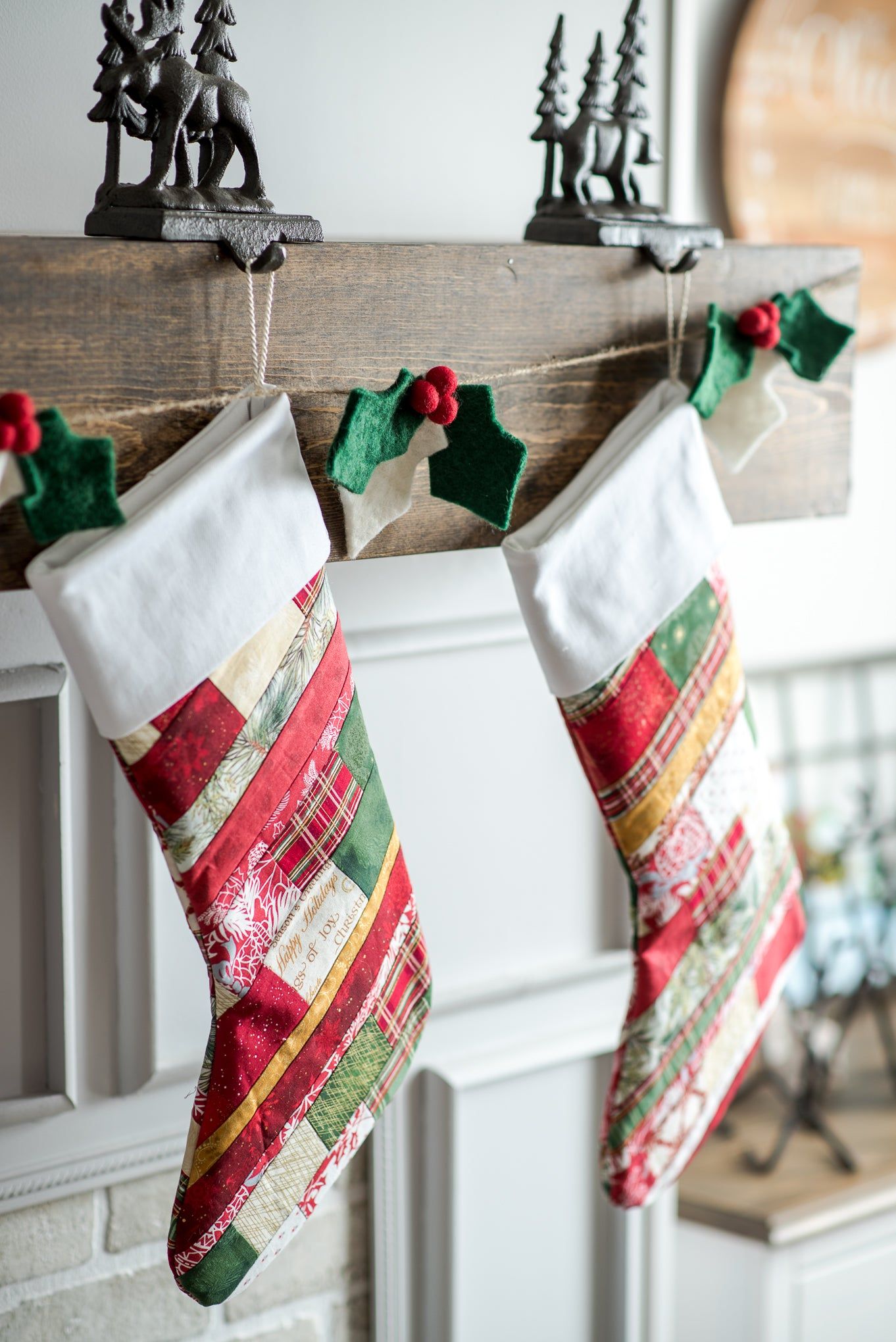 Two festive stockings with red, green, and white patchwork hang from a wooden mantel decorated with a Nivas Christmas Holly Garland.