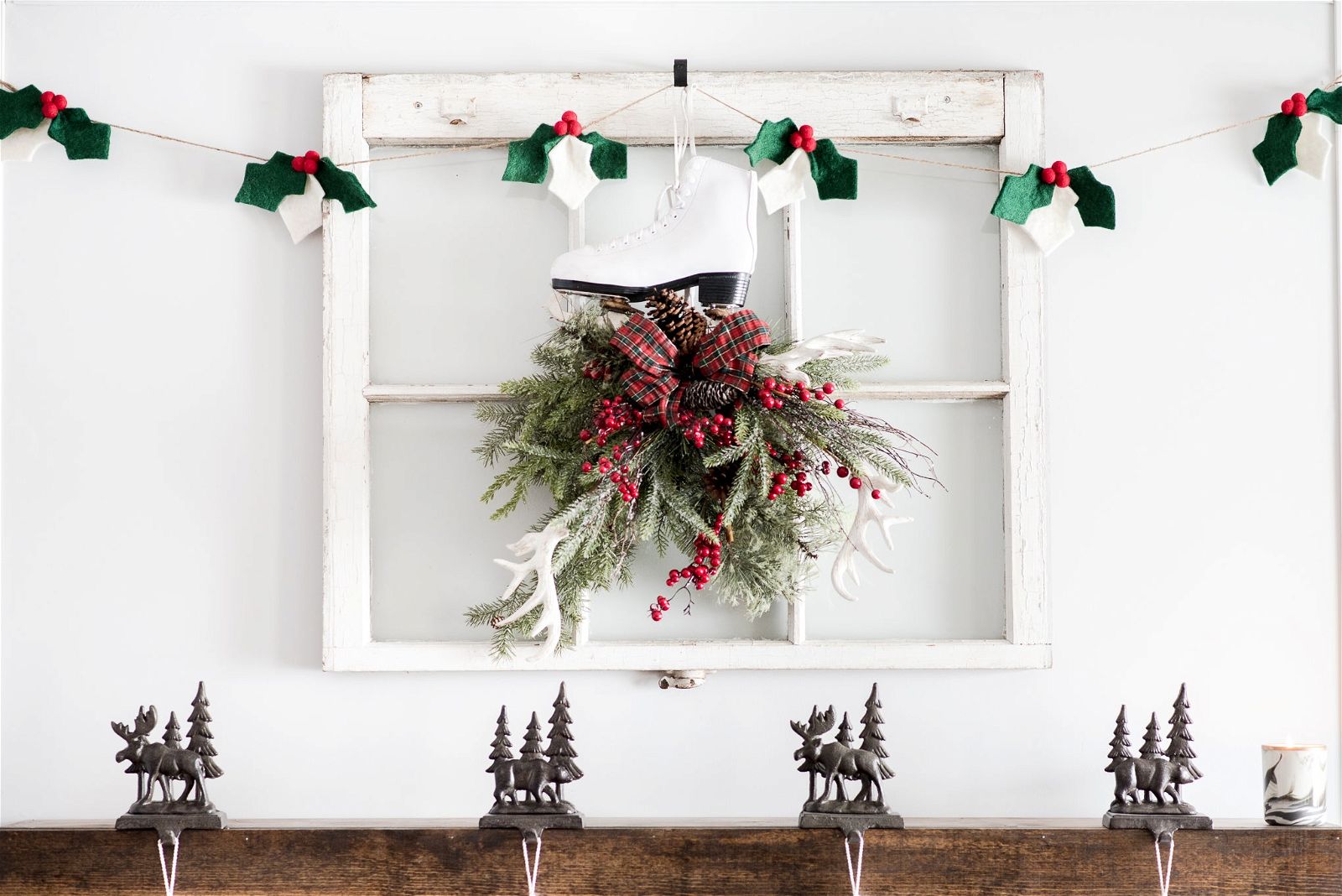 Festive decor featuring Nivas Christmas Holly Garland with a window frame displaying a white ice skate, greenery, and berries. Below, wool felt reindeer and tree figures adorn a wooden shelf.