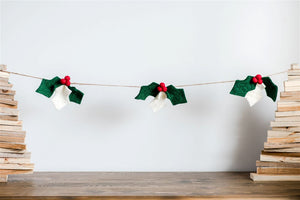 The Nivas Christmas Holly Garland, featuring green wool felt leaves, white felt bells, and red berries, elegantly drapes between two book stacks on a wooden surface, reminiscent of classic Christmas tree decorations.