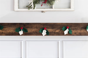 A festive mantel is adorned with a Nivas Christmas Holly Garland, showcasing green leaves and red berries, under a window decorated with pine branches, hinting at the nearby Christmas tree.