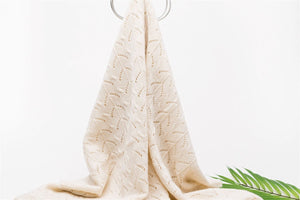 The Nivas Diamond Cashmere Baby Blanket, cream-colored and hand-knitted with a patterned design, is elegantly draped over a round metal stand with a green leaf in view.