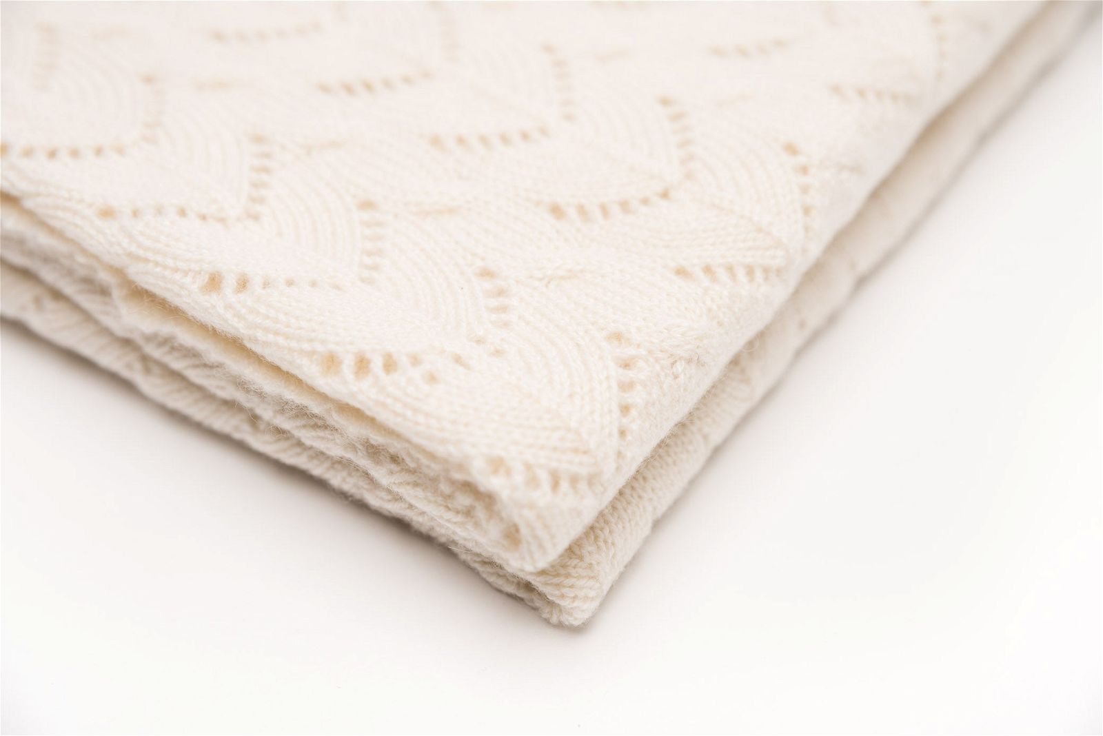 The Diamond Cashmere Baby Blanket by Nivas is a folded, cream-colored knit blanket with a subtle zigzag pattern, hand-knitted from pure cashmere.