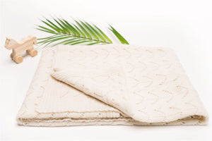 The Diamond Cashmere Baby Blanket by Nivas, cream-colored and hand-knitted, is folded on a white surface alongside a wooden horse toy and a green palm leaf.