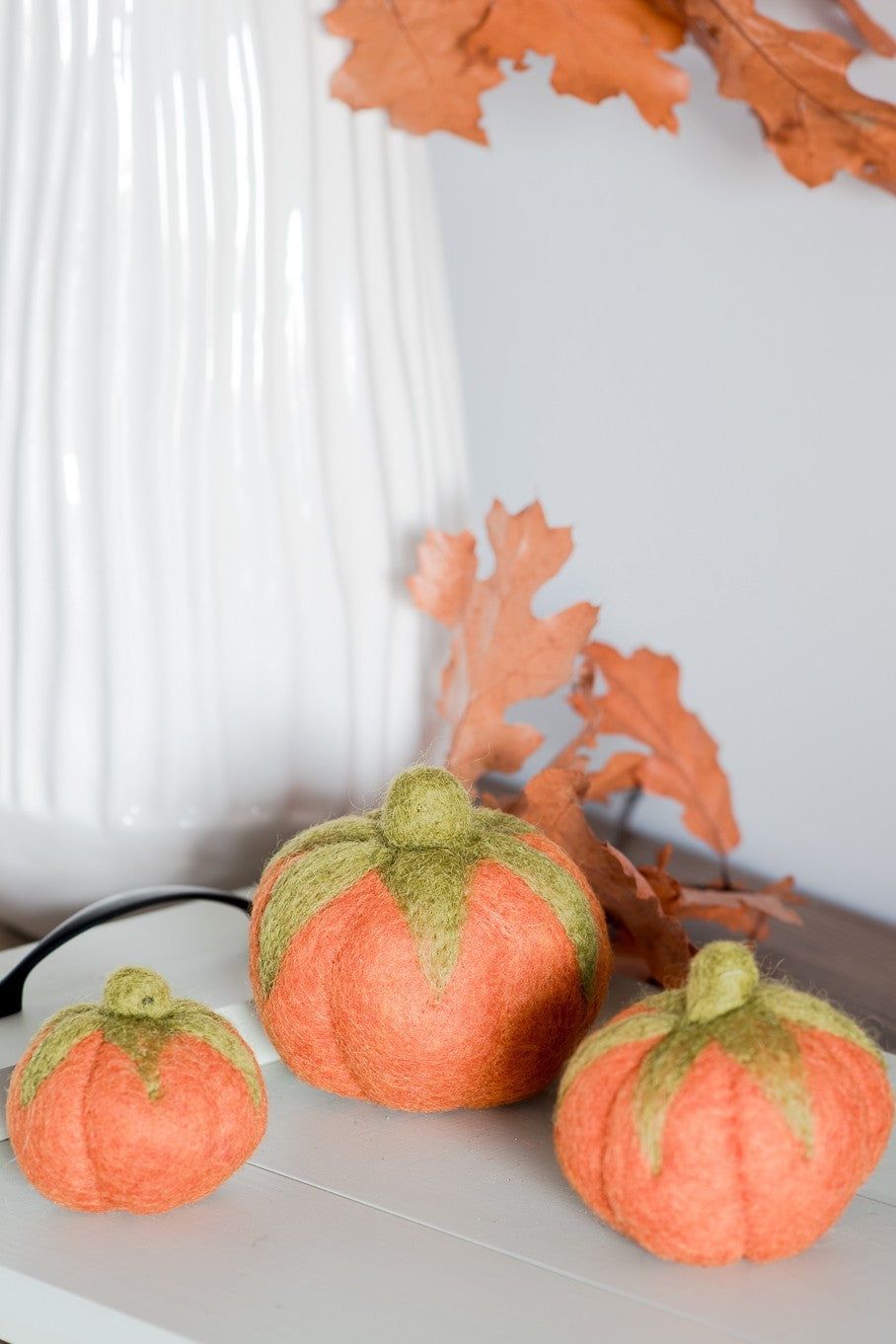 Nivas Faux Pumpkins in a trio of sizes are artistically arranged near a white vase with dried orange leaves, crafting cozy fall vibes ideal for a table centerpiece.