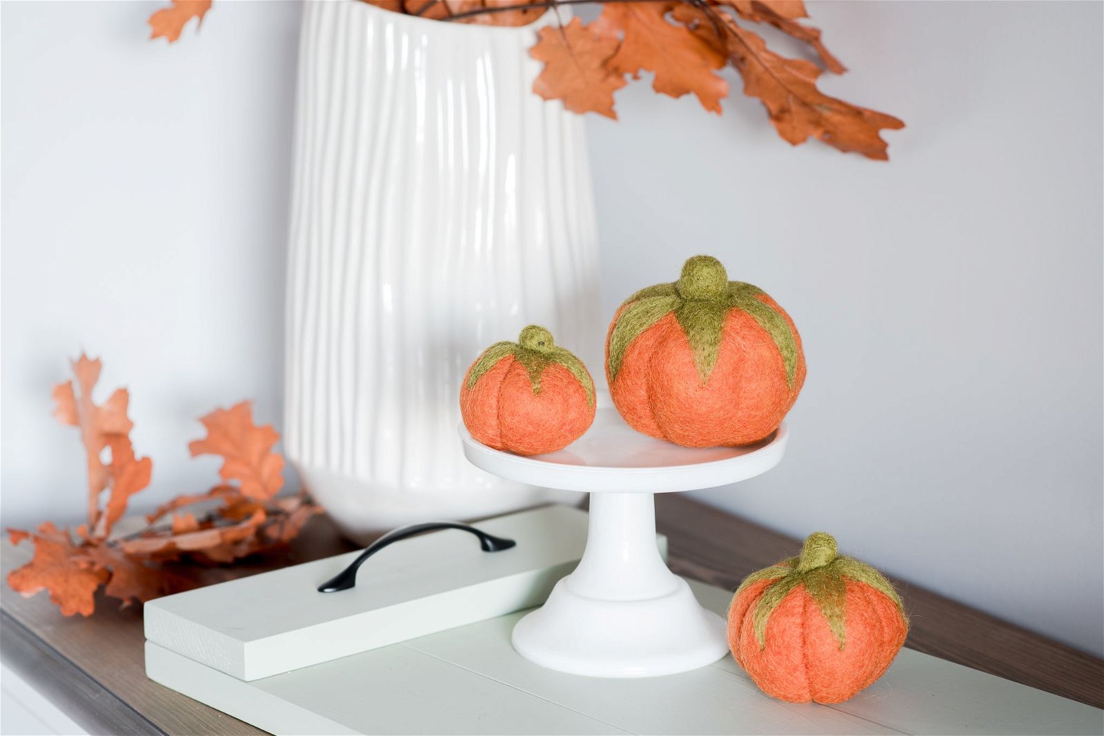 Three Nivas Faux Pumpkins with green tops are arranged as a table centerpiece on a white stand, surrounded by dry autumn leaves and complemented by a white vase, creating cozy fall vibes.