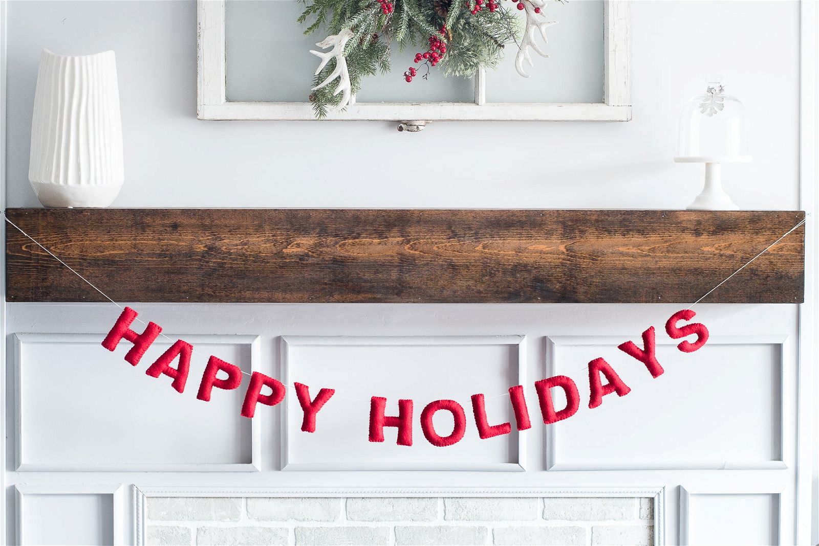 The Happy Holidays Garland (Red) by Nivas decorates a wooden mantel with festive charm. A white vase and cake stand complement the scene, enhanced by greenery and berries for delightful holiday decor.