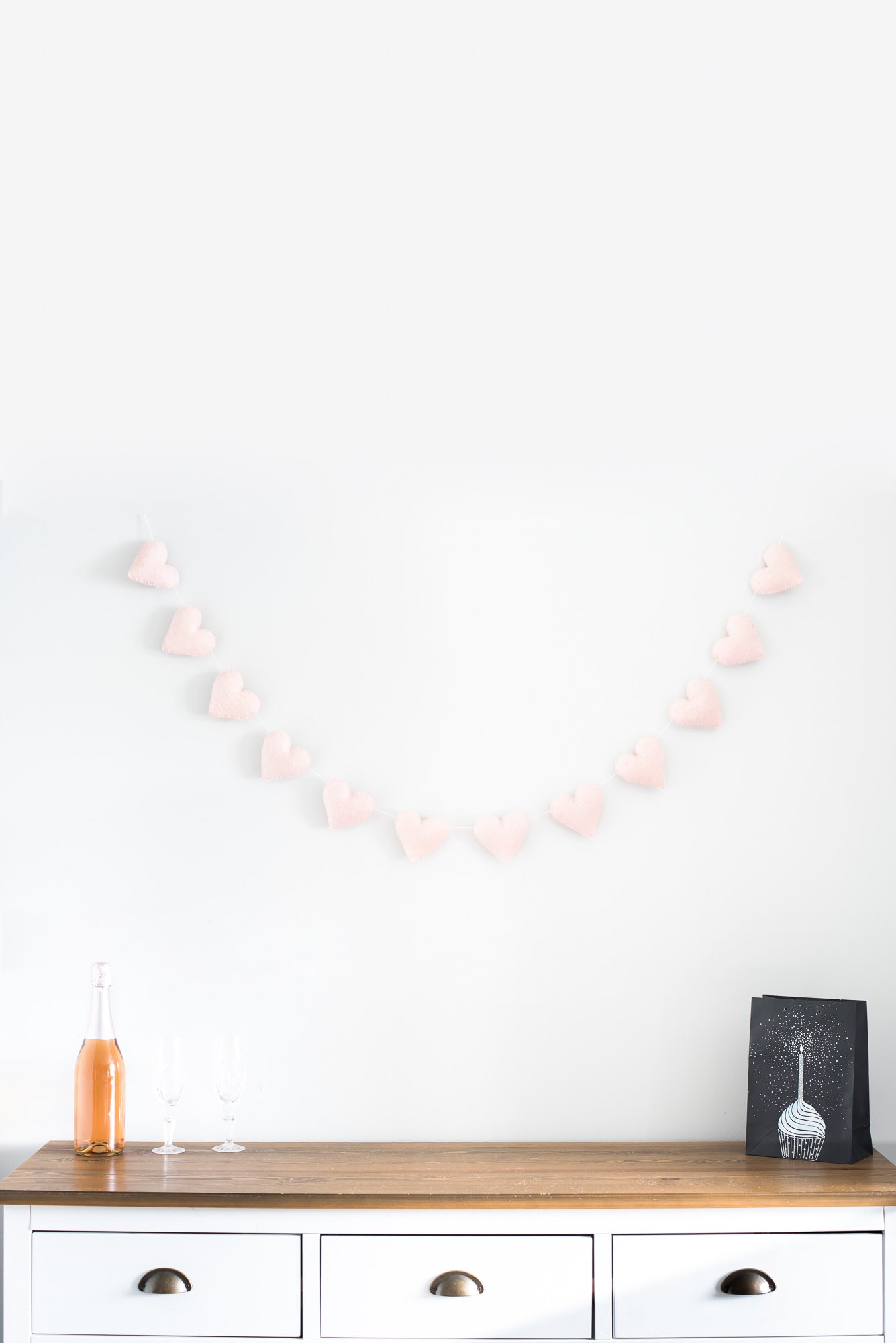 A wooden sideboard holds two empty glasses and a bottle on the left. Above, a Nivas Heart Garland (Pink) decorates the white wall. A framed picture sits to the right, completing this cozy vignette.