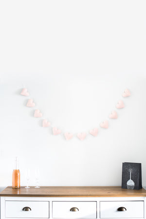 A wooden sideboard holds two empty glasses and a bottle on the left. Above, a Nivas Heart Garland (Pink) decorates the white wall. A framed picture sits to the right, completing this cozy vignette.