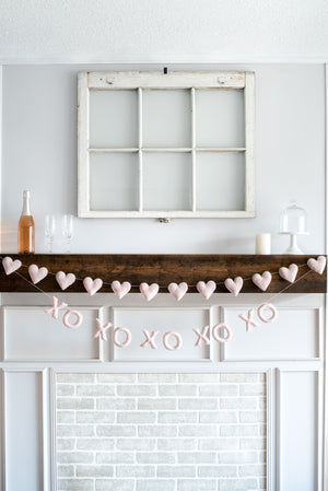 A mantel features a Nivas Heart Garland (Pink), an XO banner, two empty glasses, a bottle, candles, and a decorative window frame above a white brick fireplace.