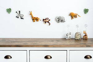 The Nivas Jungle Animal Garland, featuring a zebra, giraffe, monkey, elephant, and lion in wool felt, hangs on a white wall above a wooden dresser with three animal figurines—ideal for your safari adventure decor.