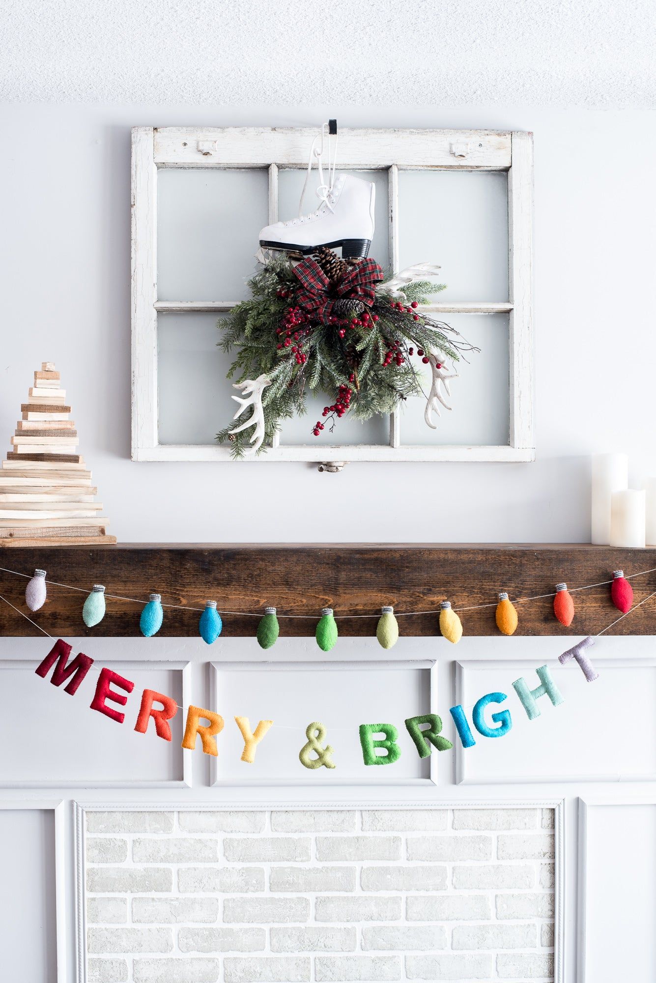 A festive mantel exudes holiday cheer with a wooden tree, candles, and Nivass Merry and Bright Garland. Above, a window frame showcases a wreath with an ice skate and pine branches, while the garland adds charm to the scene.