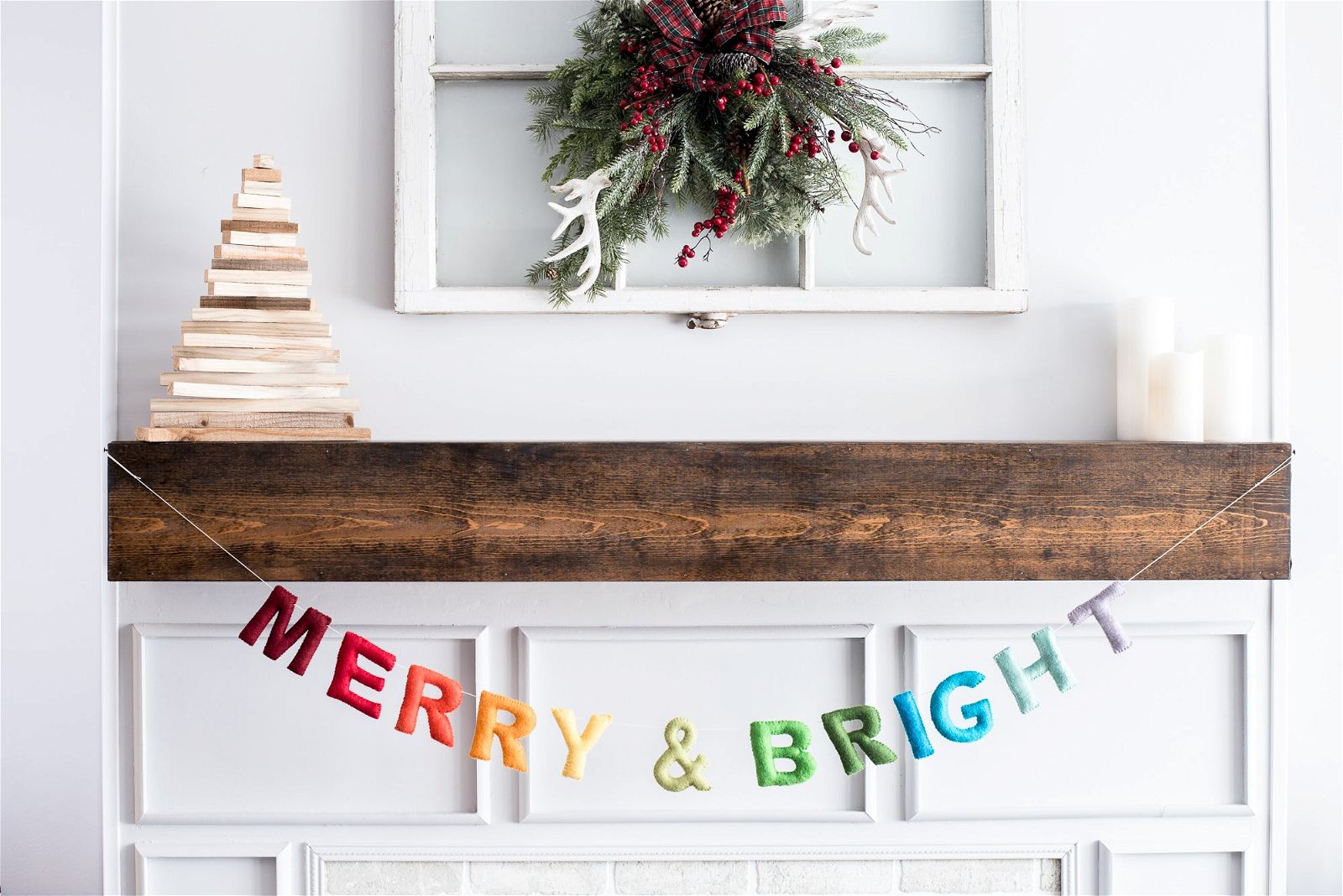 A mantel exudes holiday cheer with Nivass rainbow Merry and Bright Garland, a wooden Christmas tree, a berry wreath, and three flickering candles.