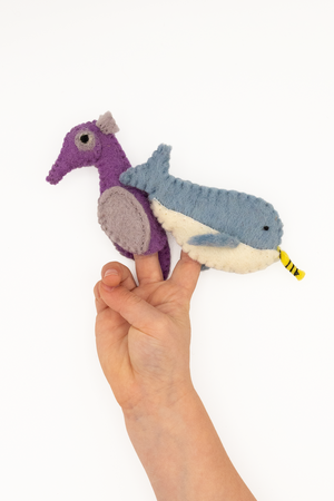 A hand displays the Nivas Narwhal & Seahorse Finger Puppet Set, crafted from wool felt and featuring a plush blue whale and purple bird against a white background.