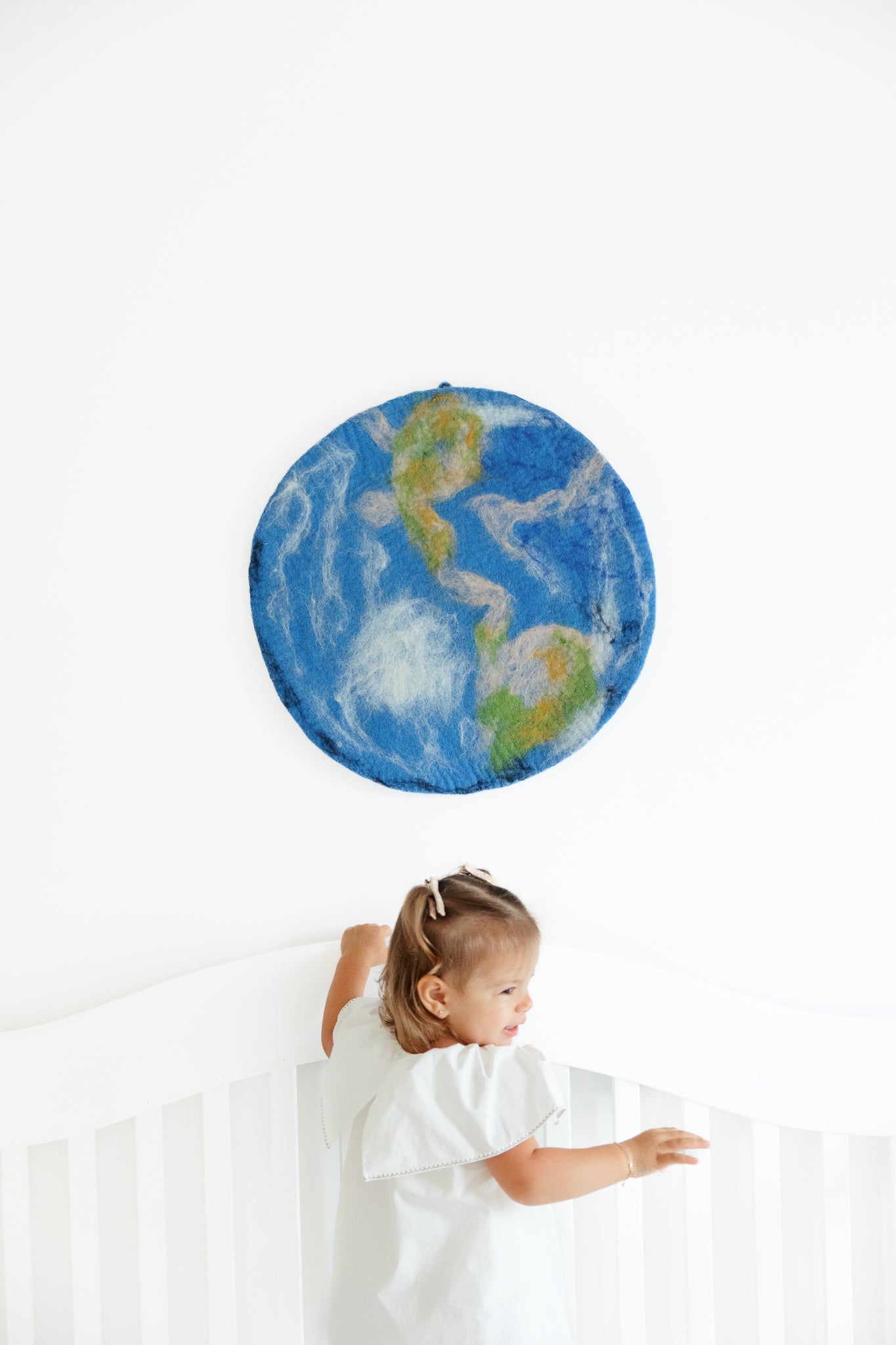 A child stands in a white crib, looking up at the Nivas Earth Wall Art, a handcrafted wool piece inspired by nature, set against a pristine white wall.