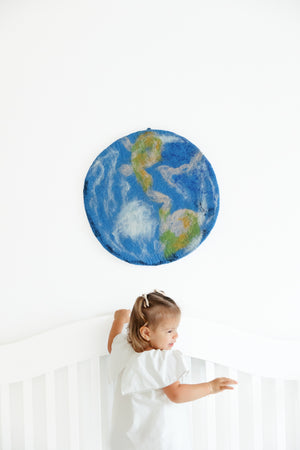 A child stands in a white crib, looking up at the Nivas Earth Wall Art, a handcrafted wool piece inspired by nature, set against a pristine white wall.