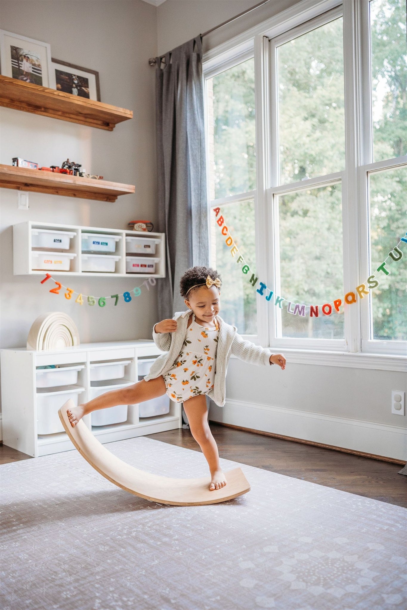 A child balances on a wooden board in a playroom, surrounded by vibrant shelves with the Nivas Alphabet & Numbers Banner (Rainbow) hanging cheerfully above, adding to the lively atmosphere.