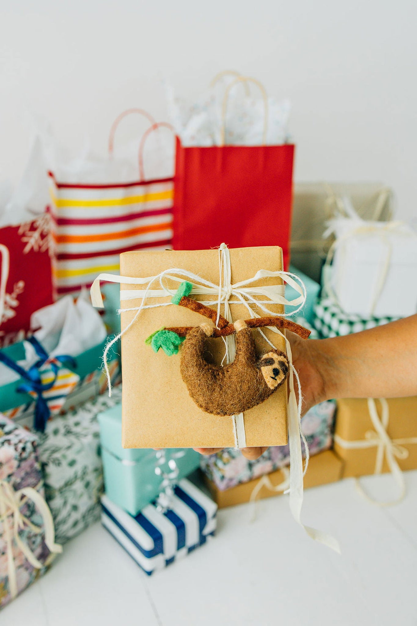 A hand holds a wrapped gift featuring a Nivas Sloth Ornament, set amid colorful holiday décor with striking gift bags and boxes.