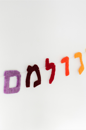 The Nivas Hebrew Garland features vibrant, rainbow-colored wool felt letters on a string, creating an eye-catching display against a white background.